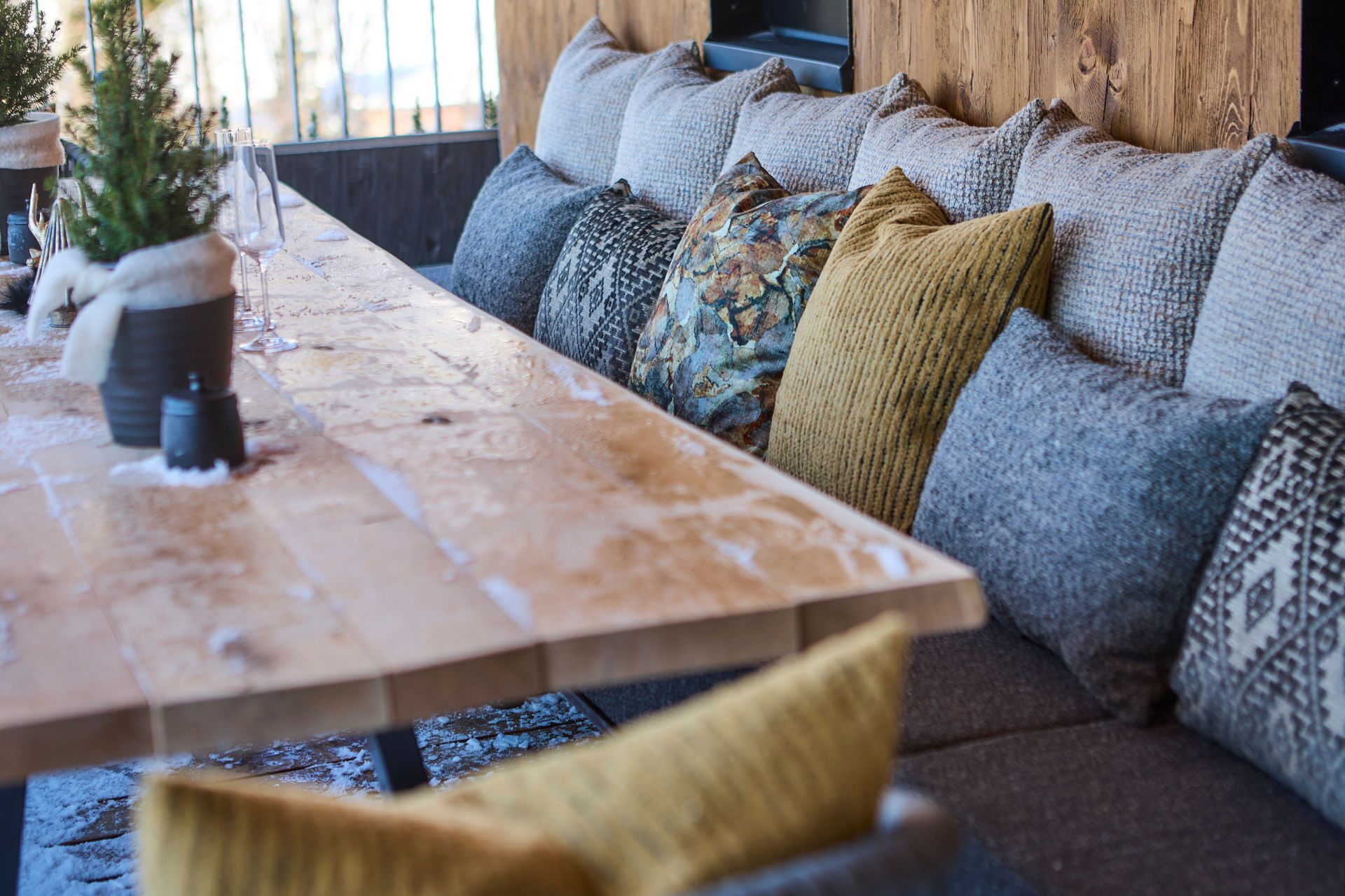 A long wooden table with a candle and glasses, a small potted plant, and a sofa with various patterned cushions.