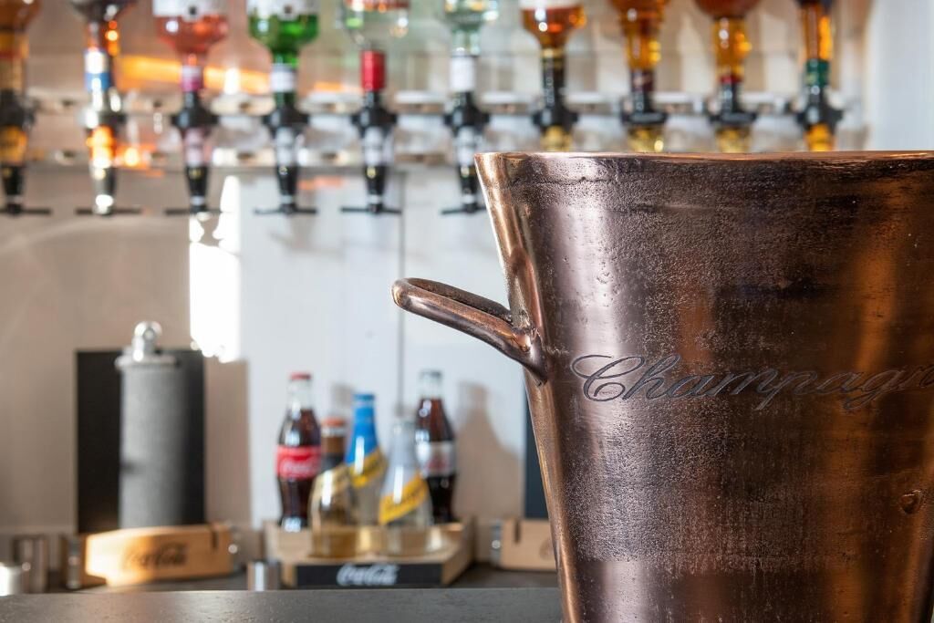 Un vaso de cobre con la palabra 'Champagne' grabada, sobre una barra con varias botellas y copas al fondo.