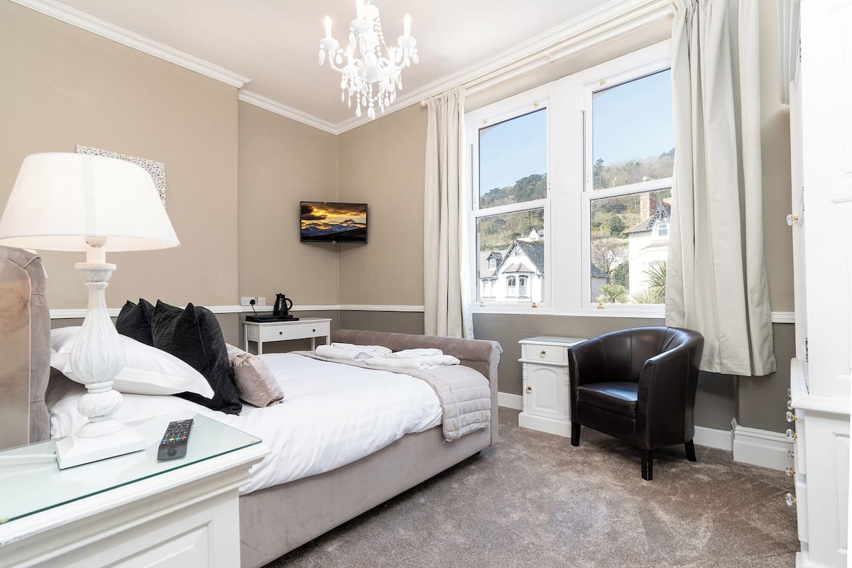 A bedroom with a bed, bedside tables, a TV, a chair, and a large window with a view of buildings.