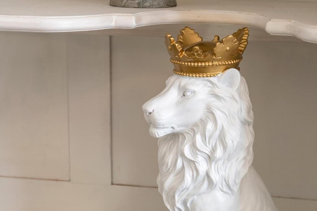 Une sculpture représentant la tête d’un lion blanc, surmontée d’une couronne dorée, posée sur une étagère blanche.