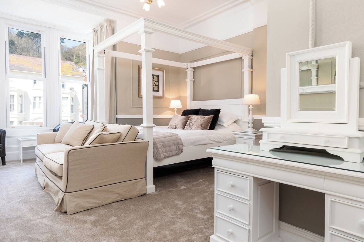 A four-poster bed with a beige and white color scheme, a desk with drawers, and a large window with a view of buildings.