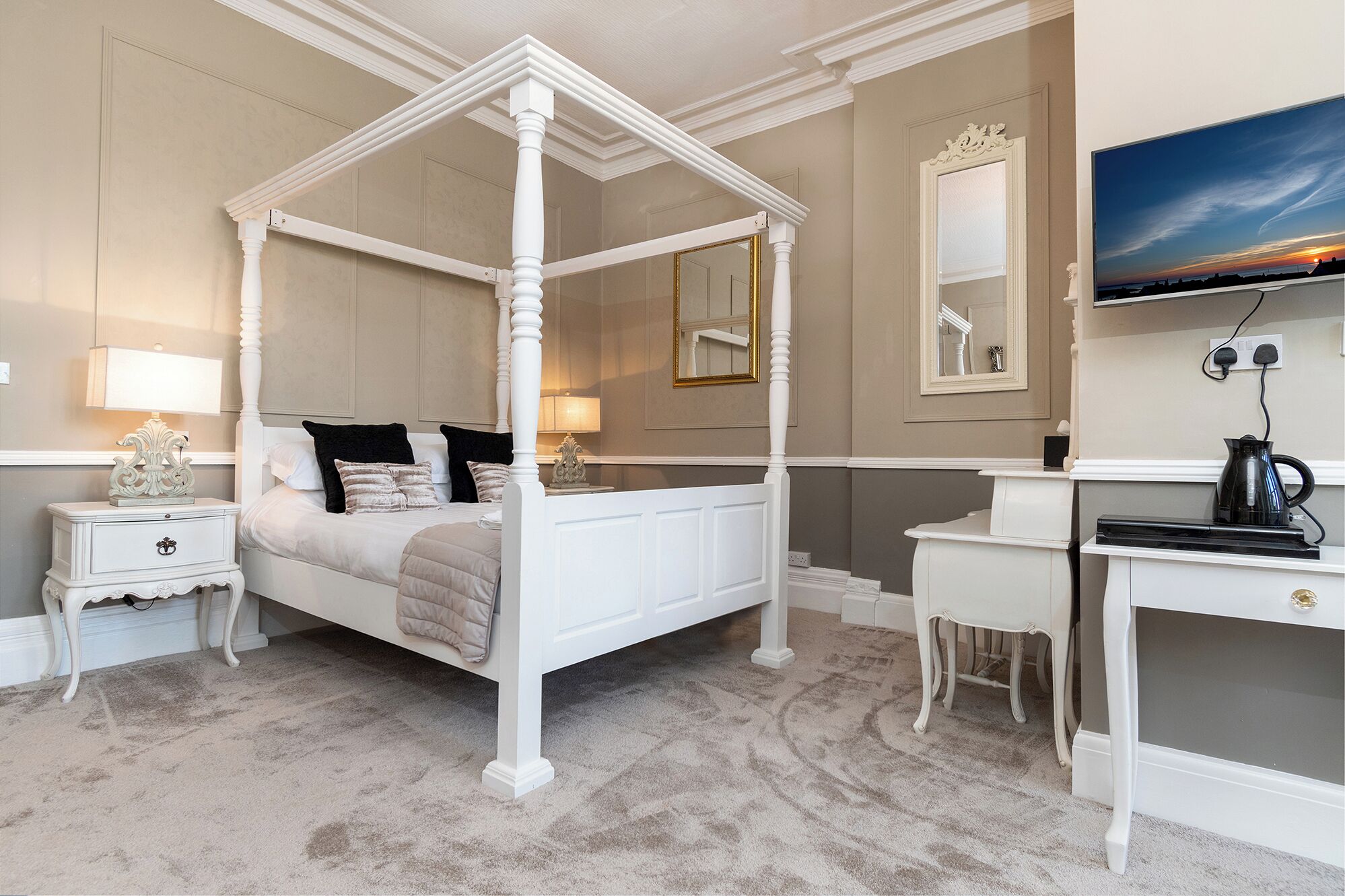 A four-poster bed with a canopy, a bedside table, a desk with a chair, a television, and a mirror on the wall.
