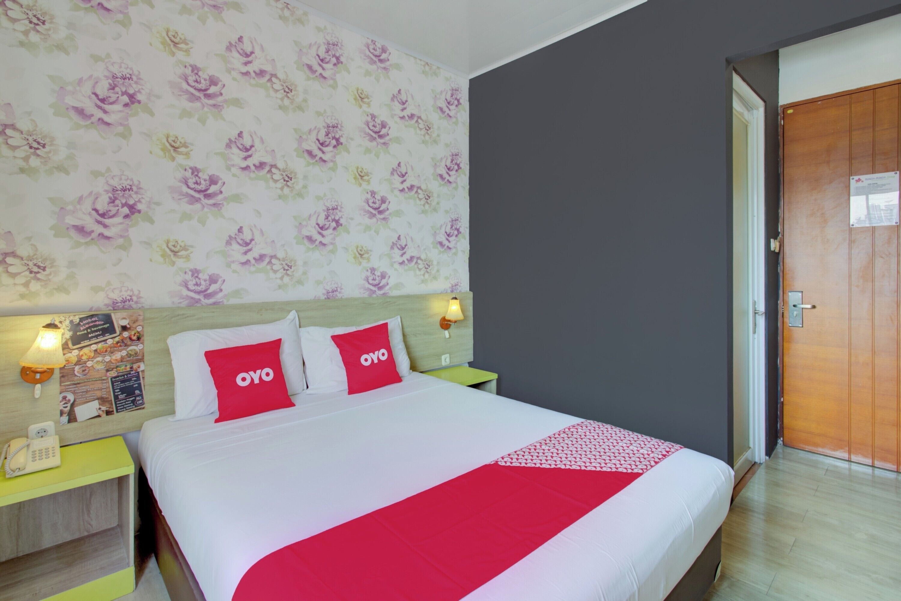 Une chambre d’hôtel avec un lit, un papier peint à motifs floraux, une porte en bois et une table de chevet avec un téléphone et une lampe.