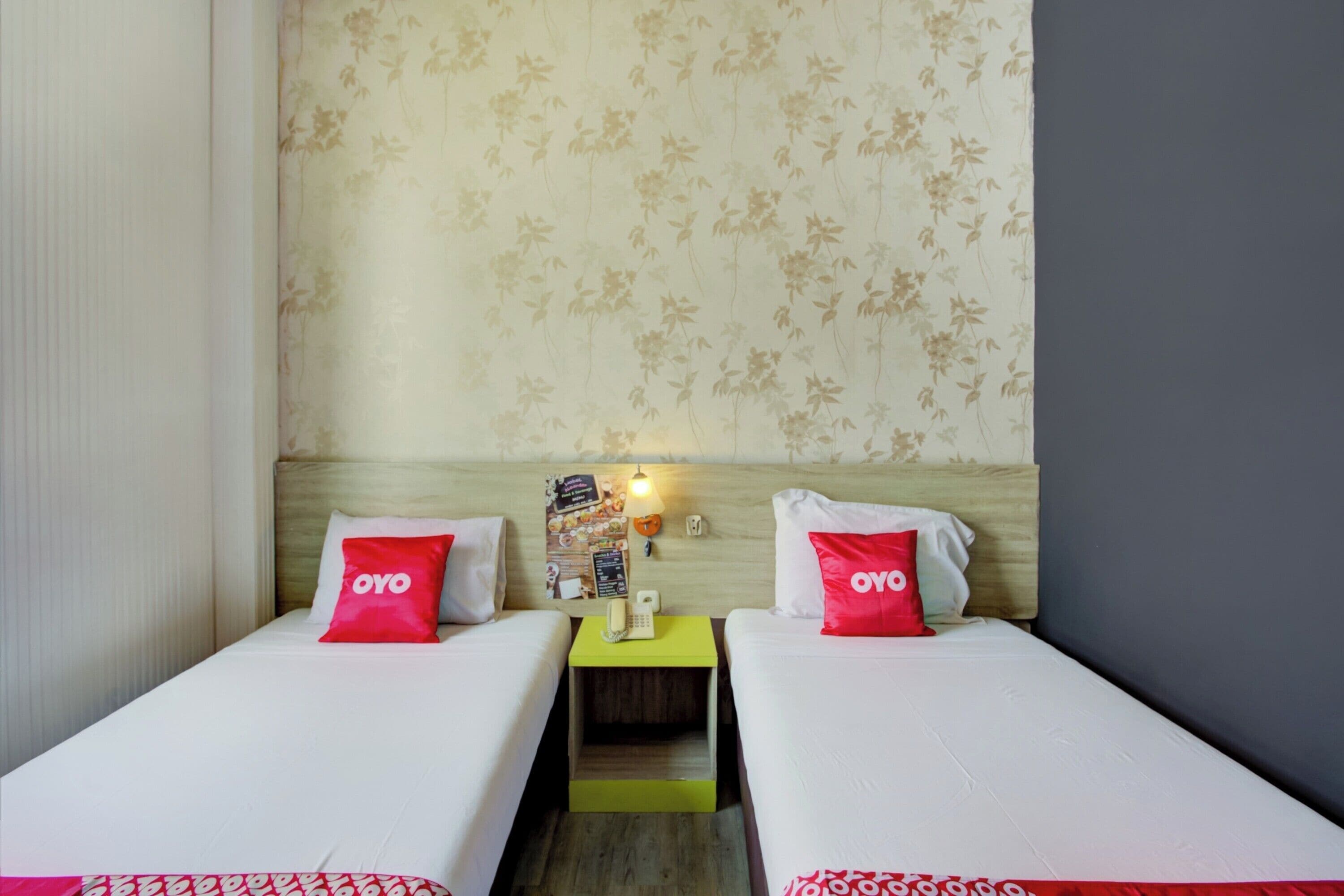 Et hotelværelse med to senge, hver med en pude mærket "OYO". Et lille bord placeret mellem sengene har en telefon og en menu. Værelset har tapet med blomstermønster og en lysefarvet sengegavl.