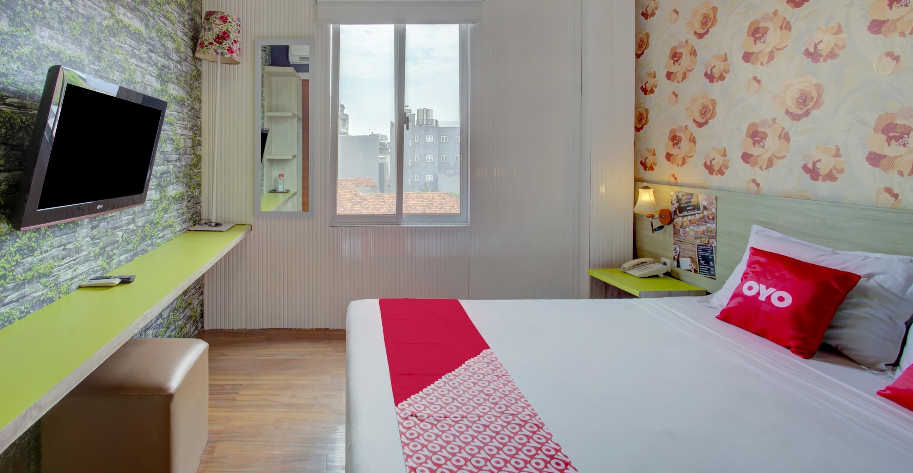A hotel room with a bed, a desk, a television, a window, and a floral wallpaper.