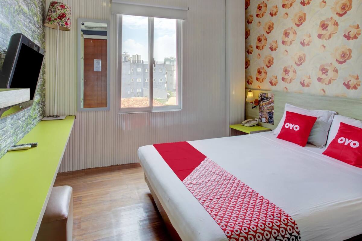 A hotel room with a bed, a desk, a television, a window, and a floral wallpaper.