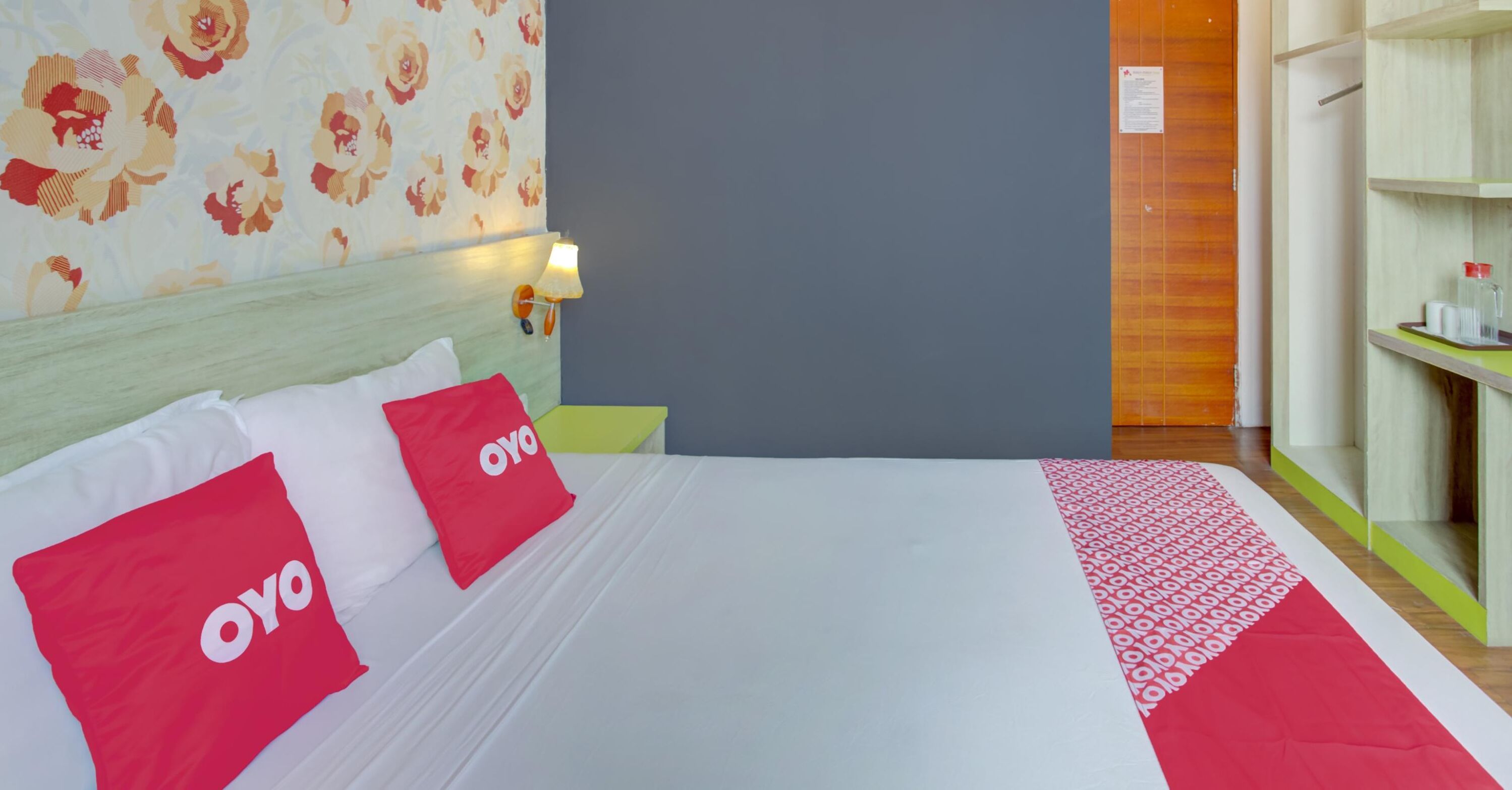 A hotel room with a bed, two red pillows with 'OYO' branding, a nightstand with a lamp, a wardrobe, and a floral wallpaper.