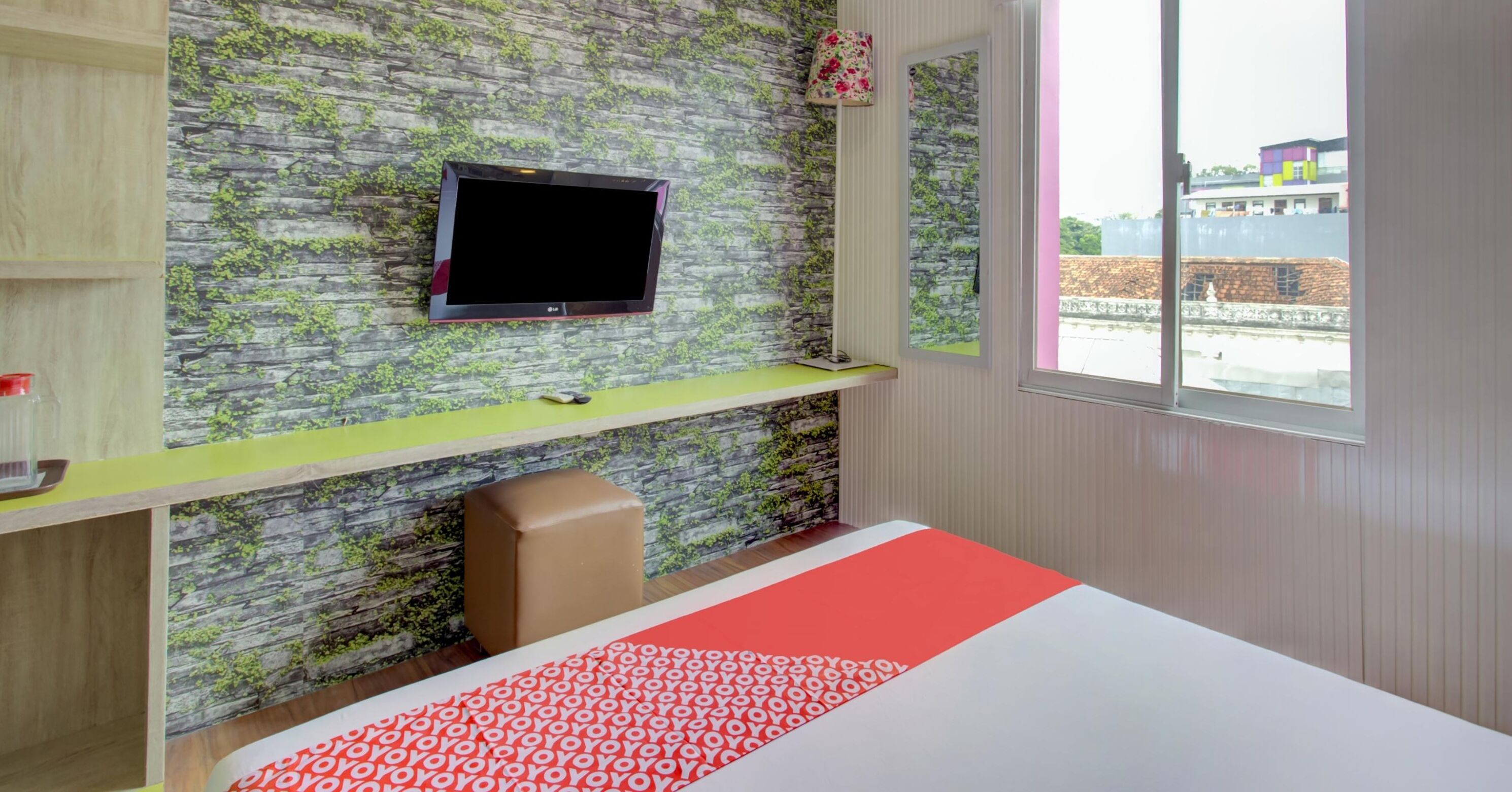 Une chambre d’hôtel moderne avec un mur décoré d’un motif en pierre, un téléviseur à écran plat fixé au mur, un bureau vert, un siège marron rembourré et une grande fenêtre donnant sur des immeubles.