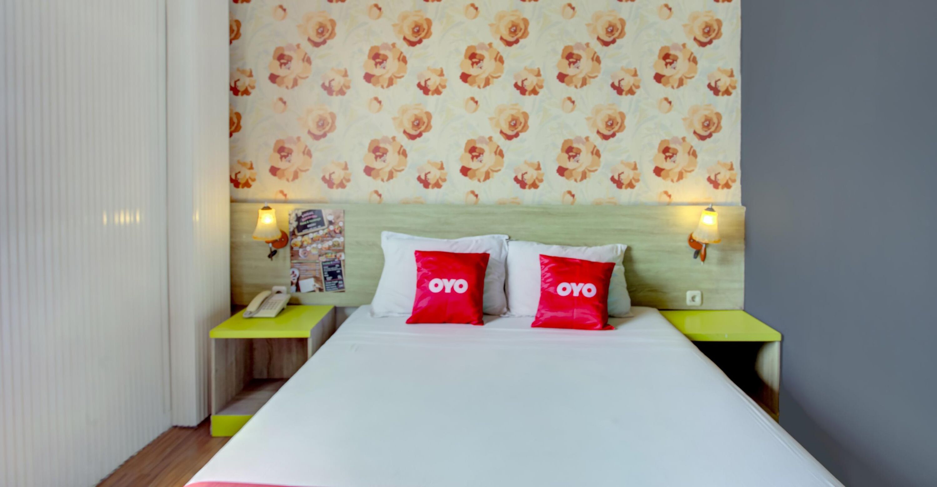 A hotel room with a bed, two nightstands, a floral wallpaper, and two red pillows with the word "OYO".