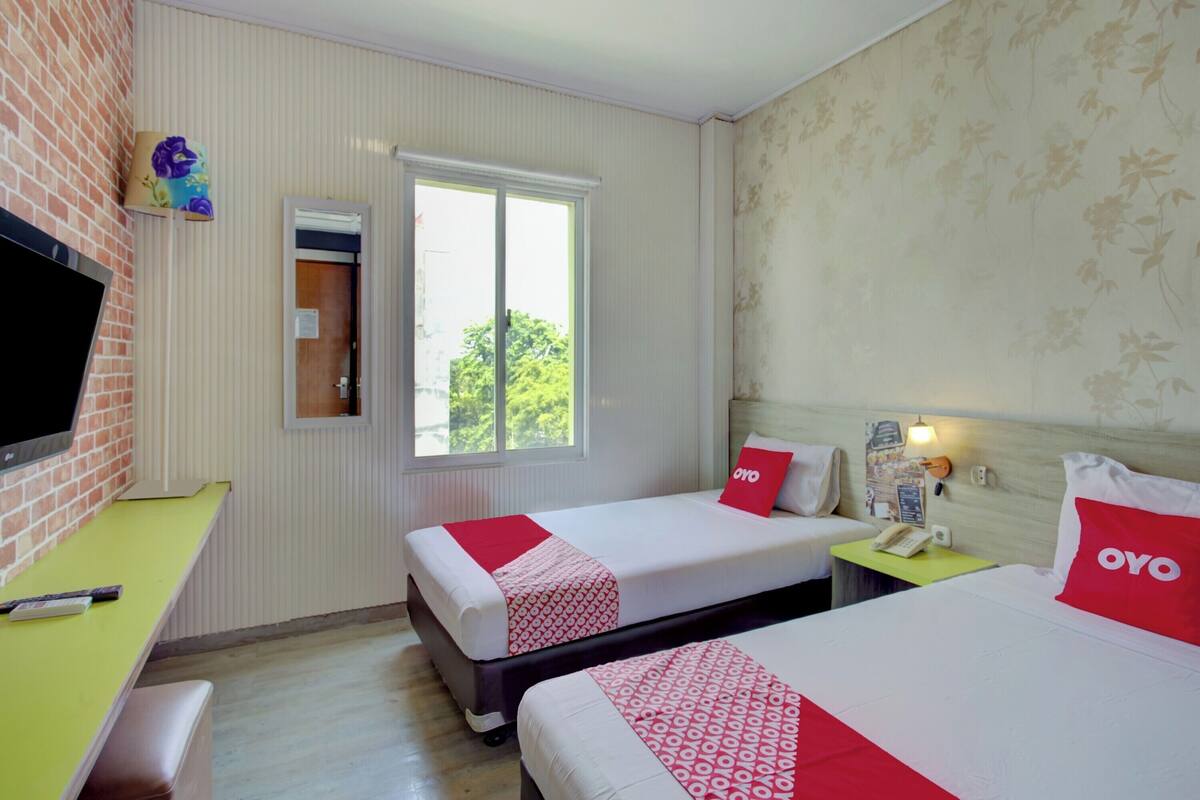 A hotel room with two beds, a TV, a desk, and a window with a view of trees.