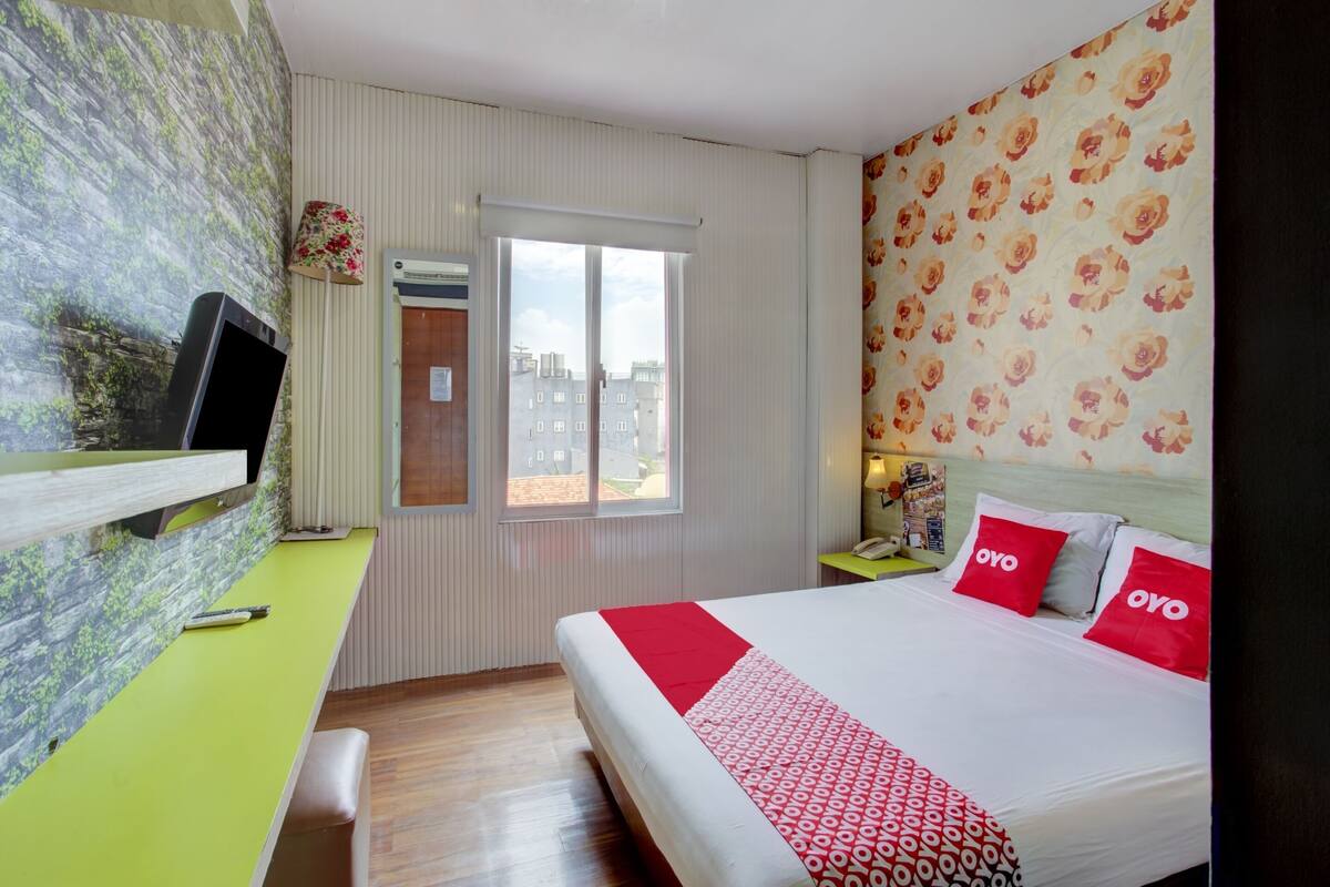 A hotel room with a bed, a desk, a television, a window, and a patterned wall.