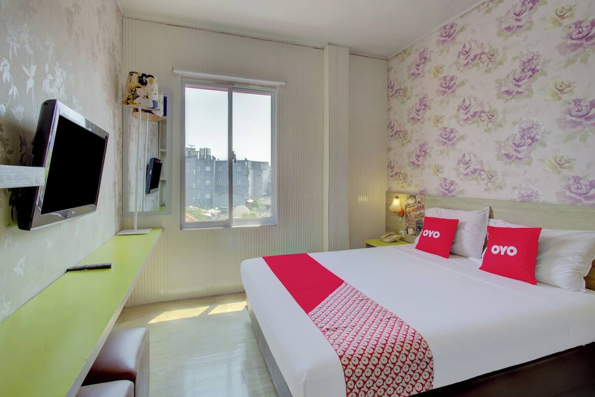 A hotel room with a bed, a desk, a TV, and a window with a view of buildings.