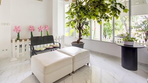 A modern room with a white marble floor, a bench with cushions, a potted tree, and a view of the outdoors through large windows. - Collection O Gambir Near Masjid Istiqlal Formerly Aleander Hotel (Jakarta)