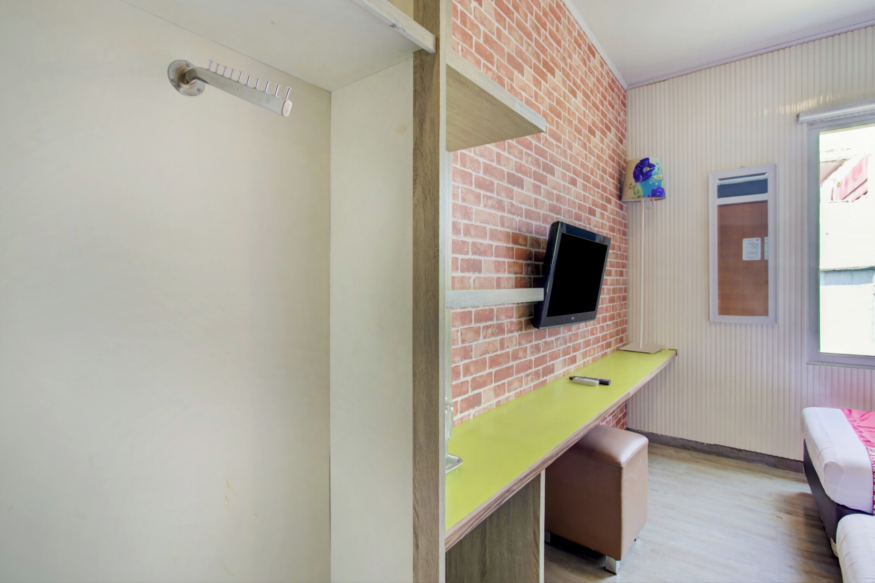 A room with a brick wall, a mounted TV, a yellow desk, a brown bench, and a window with blinds.