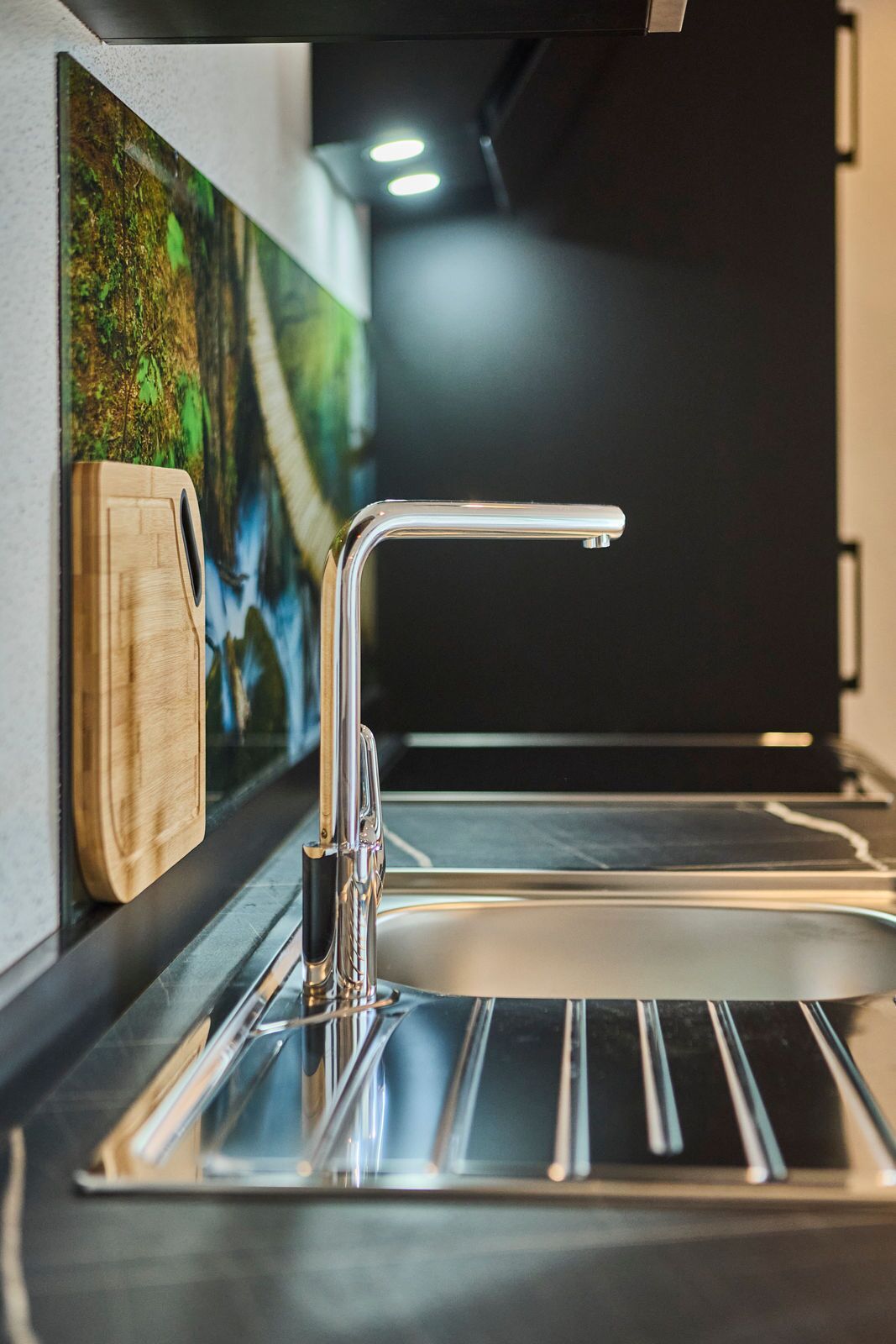 A modern kitchen with a stainless steel sink, a wooden cutting board, and a painting on the wall.