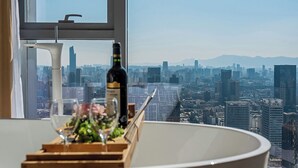 A table with a bottle and glass, a flower arrangement, and a view of a cityscape through a window. - Hotel high sky view hotel (Kunming)