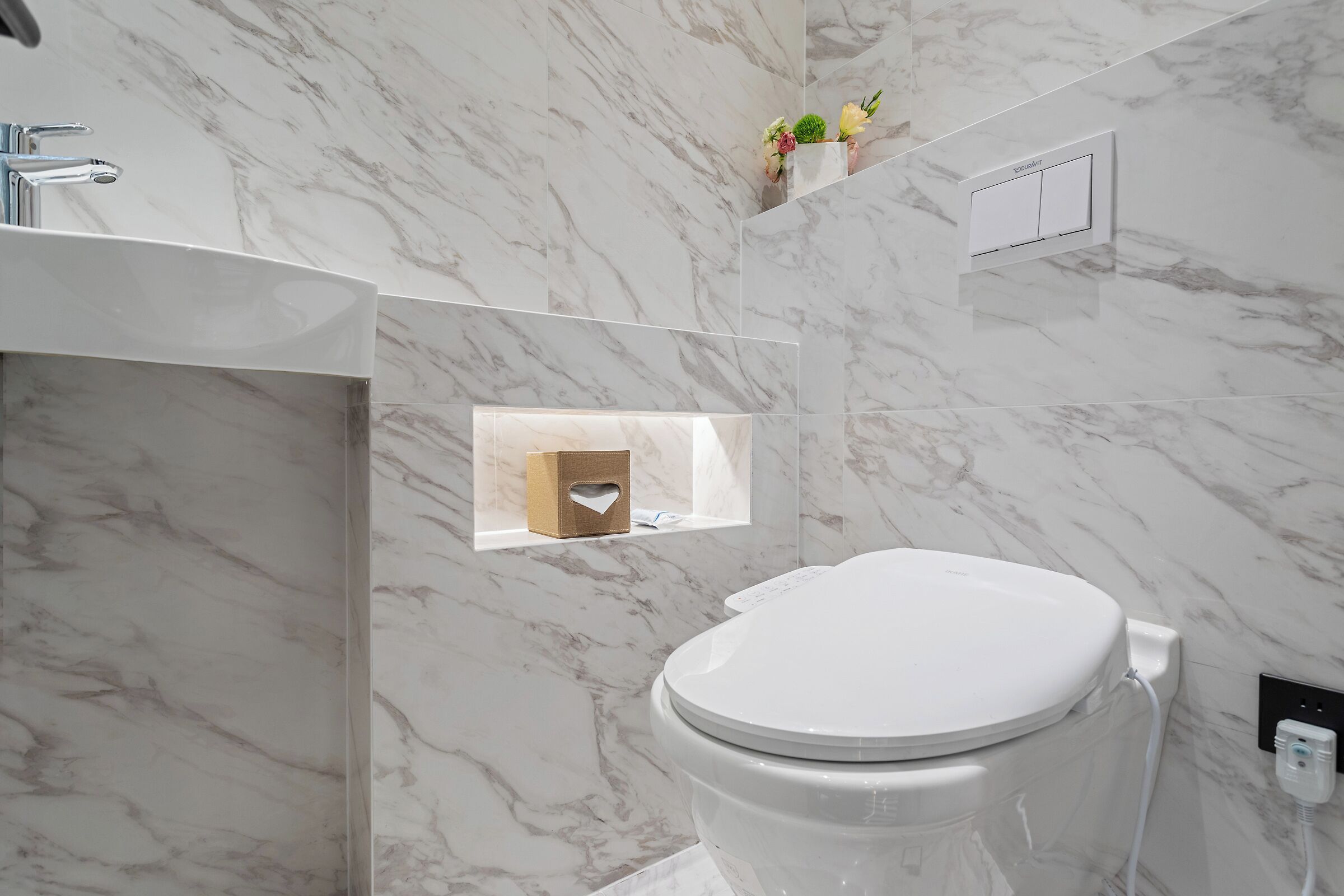 A bathroom with a marble floor, a toilet, a sink, a towel dispenser, and a small window with a tissue box.