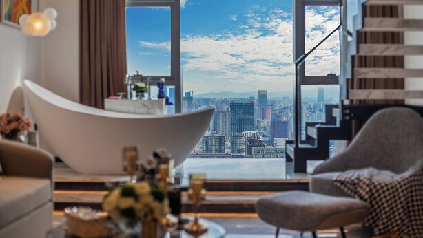 A modern living room with a large window offering a city view, a white curved sofa, and a houndstooth patterned armchair. - Hotel high sky view hotel (Kunming)