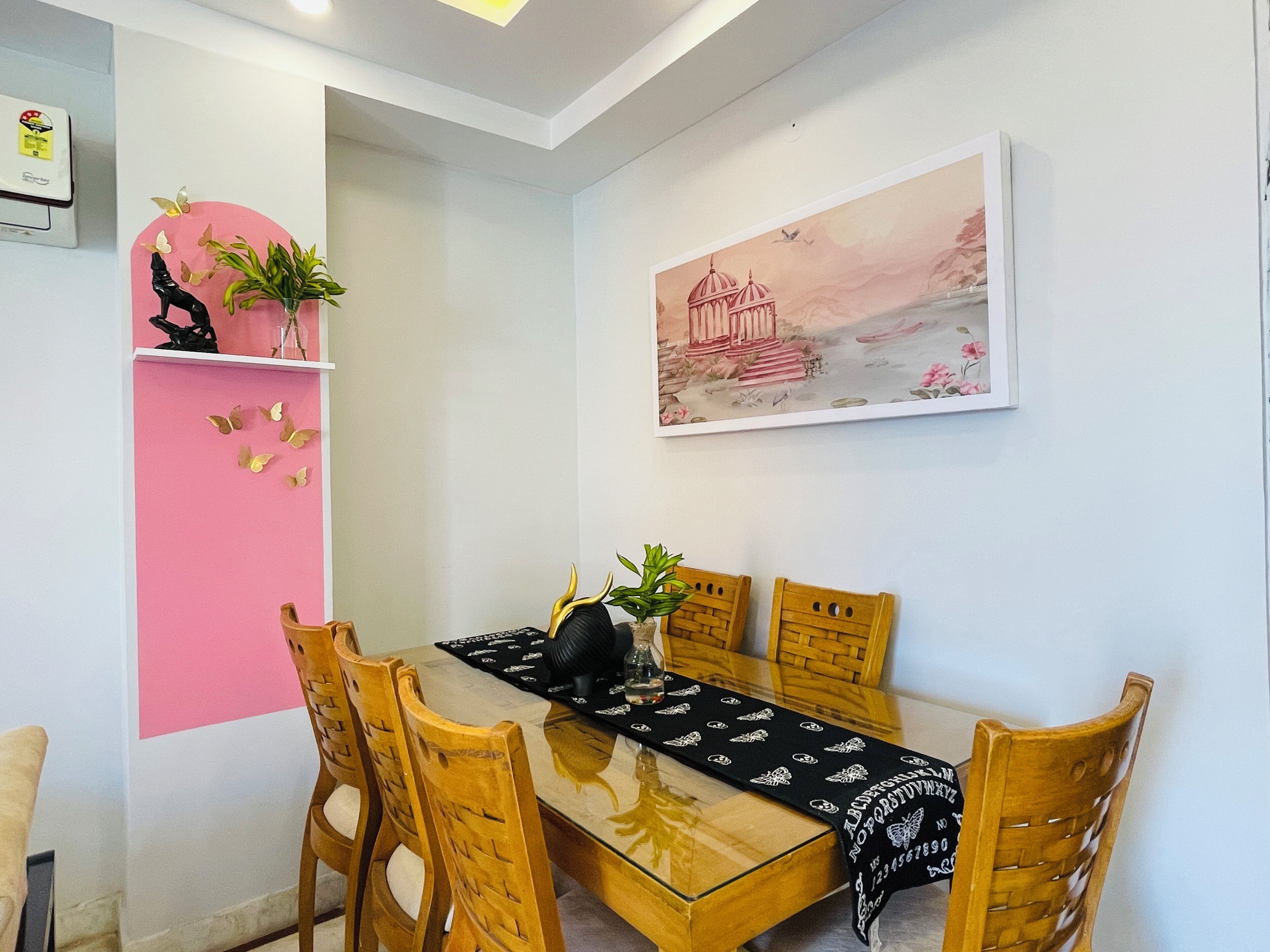 A dining area with a glass table, wooden chairs, a pink wall, and a framed picture.