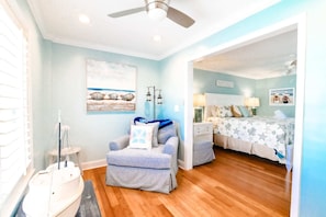 A room with a wooden floor, a ceiling fan, a bed with a starfish pattern bedspread, a chair with a blue cushion, and a wall with a seashell painting.