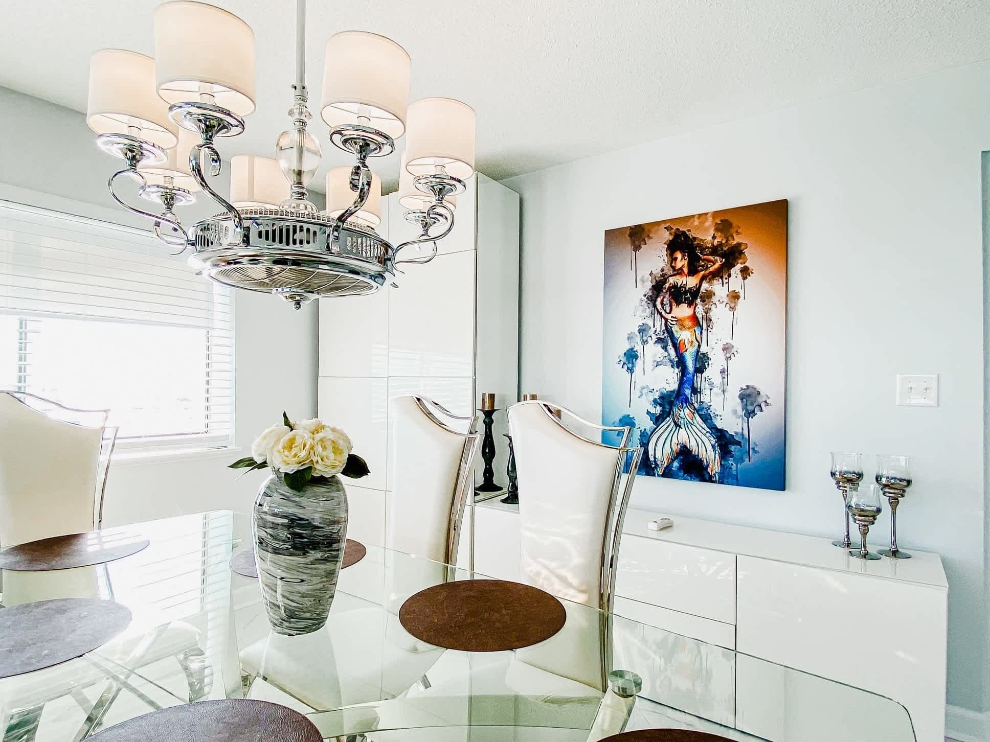 Un espace repas moderne avec une table en verre, des chaises blanches, un vase rempli de fleurs, un lustre et un tableau accroché au mur.