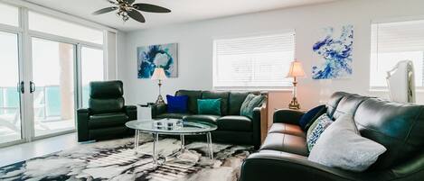 A living room with a glass coffee table, two sofas, a ceiling fan, and artwork on the walls.