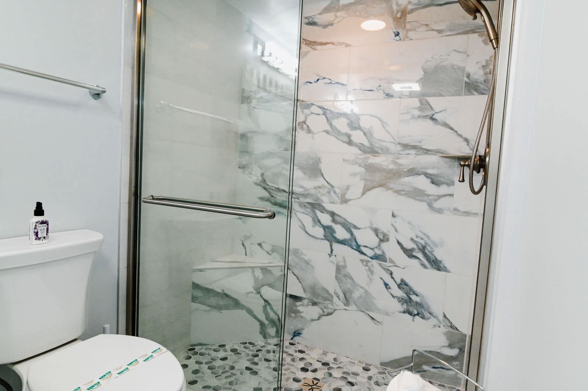 A modern bathroom with a glass shower enclosure, marble tiles, a toilet with a white lid, and a small bottle of hand sanitizer on the toilet tank.