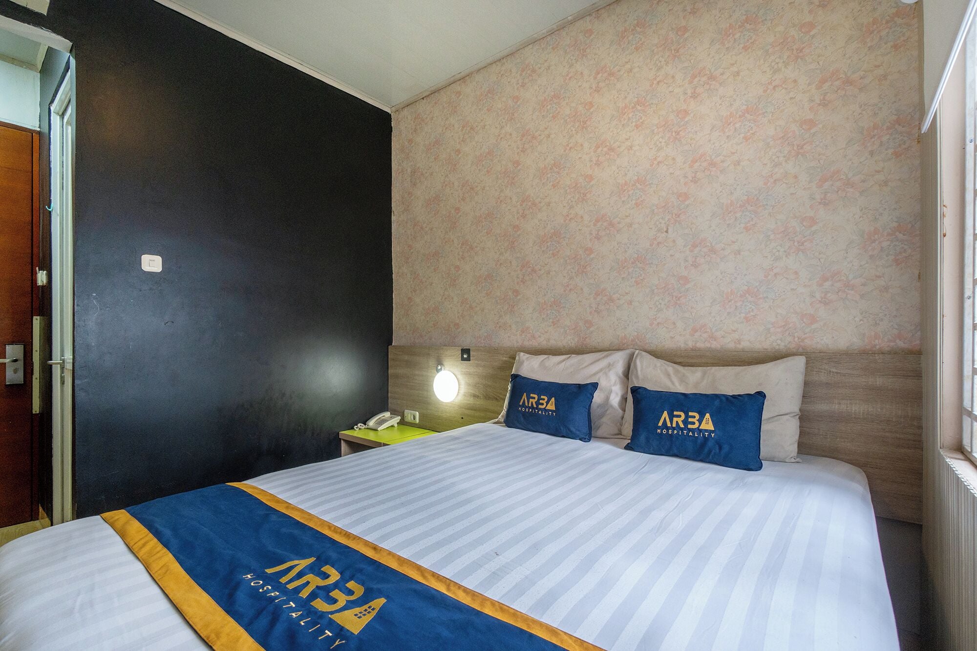 A hotel room with a bed, a nightstand, a lamp, and a floral-patterned wall.