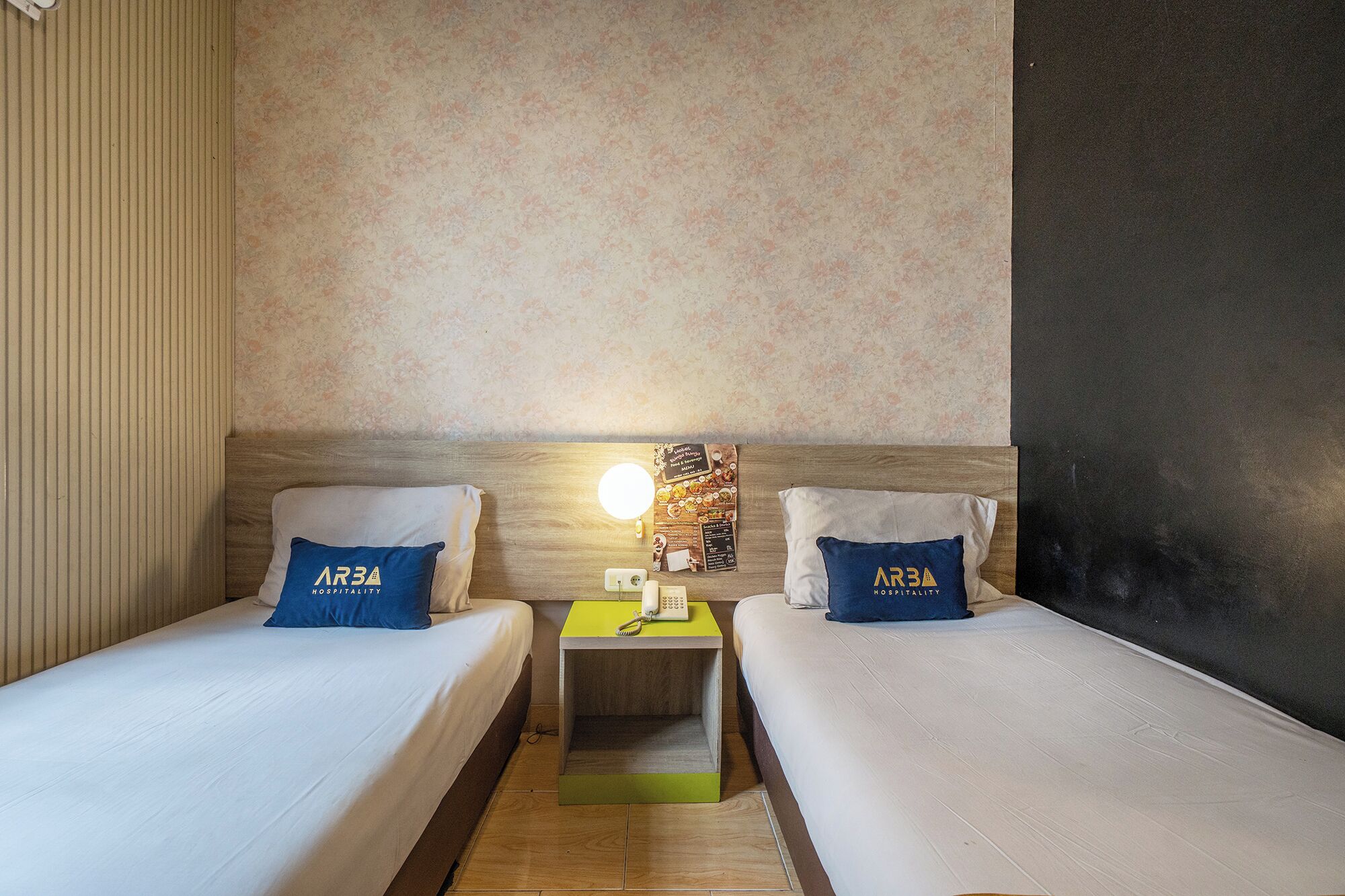 A hotel room with two beds, a nightstand with a phone, a wall clock, and a floral wallpaper.