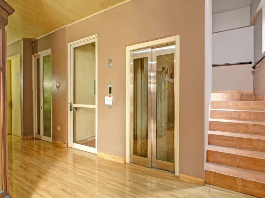 Un pasillo con suelo de madera, un ascensor de vidrio y una escalera con baldosas color beige.