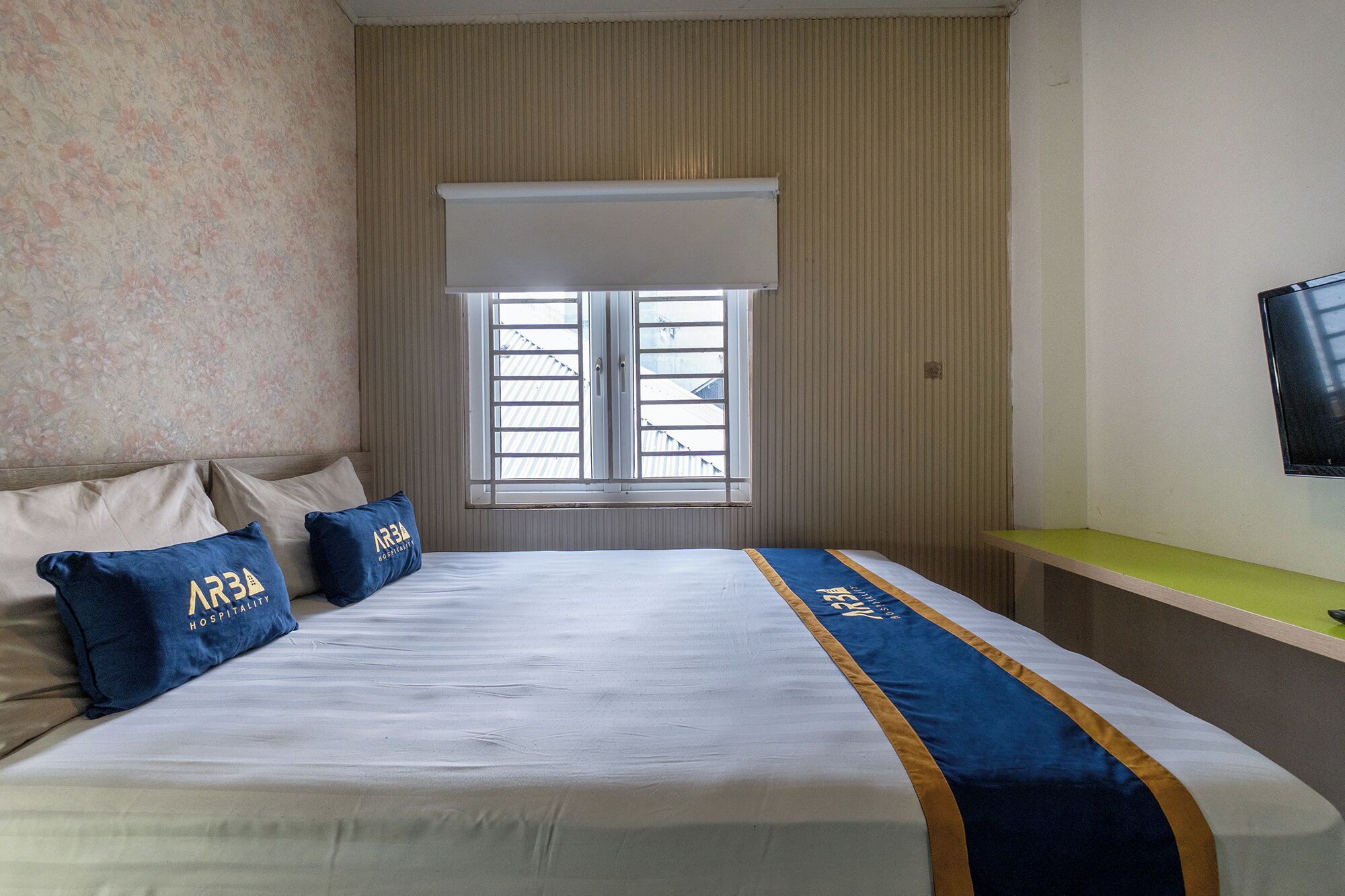 A hotel room with a bed, pillows, a window with blinds, a television on the wall, and a green bench.