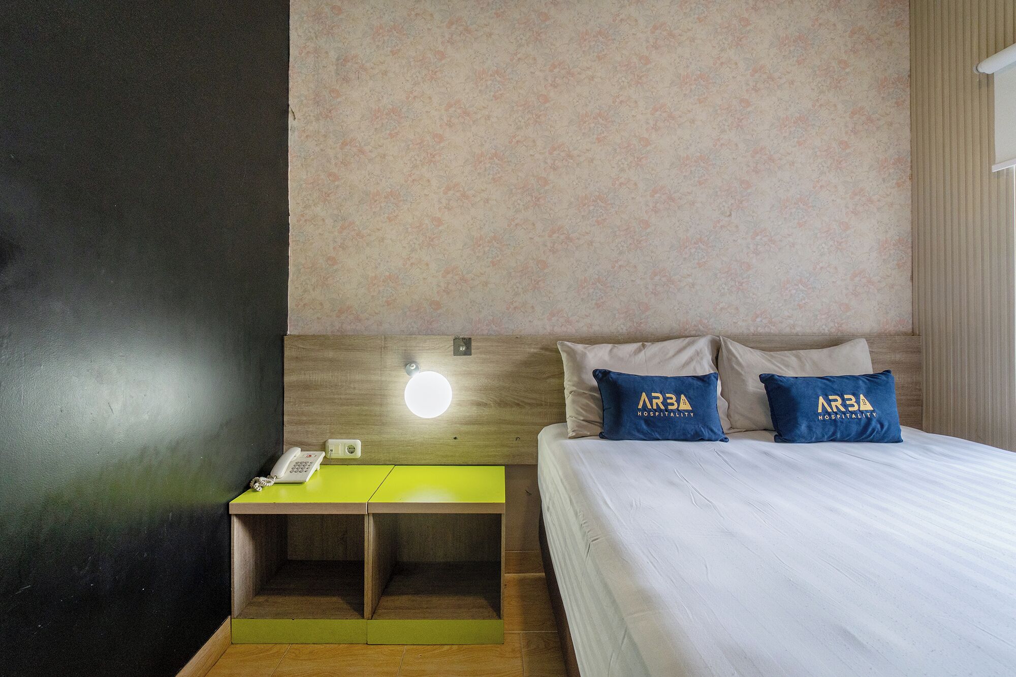 Habitación de hotel con cama, dos almohadas con el logotipo 'ARBA HOSPITALITY', mesita de noche con teléfono y luz empotrada en la pared.