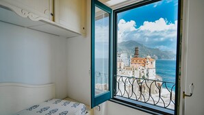 A bedroom with a bed, a window with a view of a coastal town and mountains. - Casa Blue Torricella by Elite Villas (Atrani)