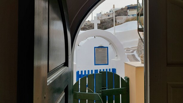 A view through an arched doorway with a white wall and blue accents, leading to a street with buildings and a gate. - Akrotiri Cosy Studio with Patio and View (Santorini)