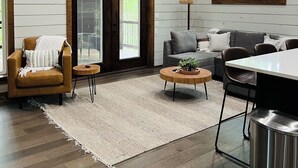 A spacious living room with a high ceiling, wooden beams, a large window, a sofa, a coffee table, and a dining area. - Eagle Forest-Sleeps 4, Hot Tub, Fireplace (Broken Bow)
