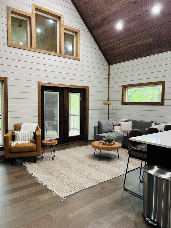 A spacious living room with a high ceiling, wooden beams, a large window, a sofa, a coffee table, and a dining area.