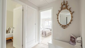 A hallway with a white door leading to a bedroom, a white dresser with a pillow, and a mirror with an ornate frame. - Windsor Charm : Spacious 4-Bed, Parking & Garden (Berkshire)