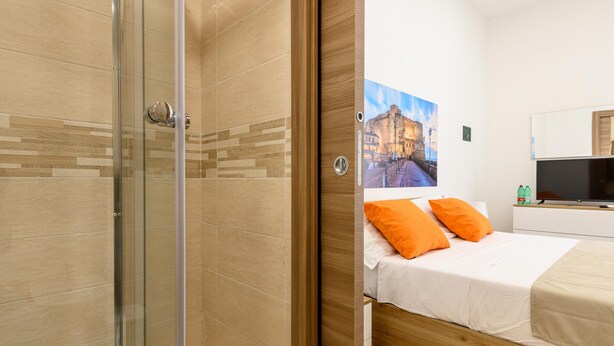 A bedroom with a bed, a wooden headboard, a painting of a castle, a TV, and a bathroom with a glass shower enclosure.