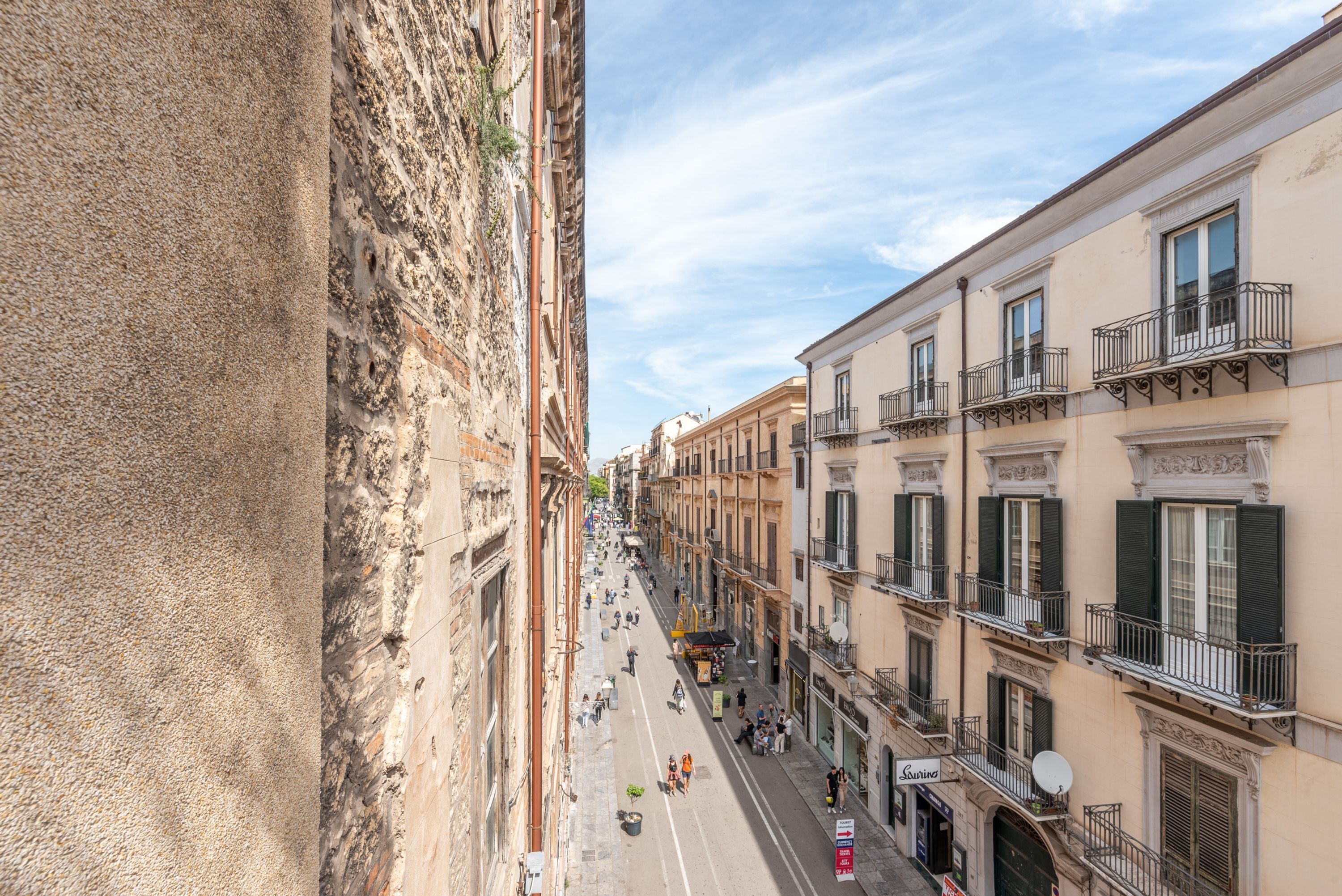Vista da rua de um edifício histórico com calçada estreita, lojas e mesas ao ar livre.