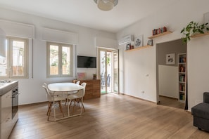 A modern kitchen with white cabinetry, a dining area with a white table and chairs, and a living area with a grey sofa.