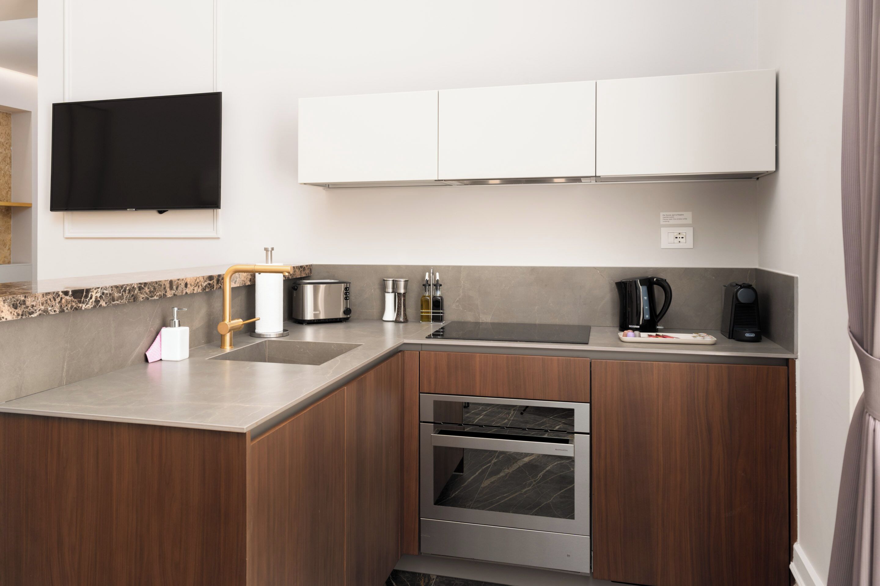 A modern kitchen with a built-in oven, a coffee maker, and a flat-screen TV mounted above the counter.
