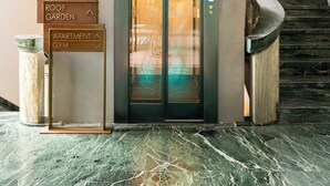 Entrada de hotel con piso de mármol, un ascensor y señalización que indica el jardín en la azotea y el gimnasio de apartamentos.