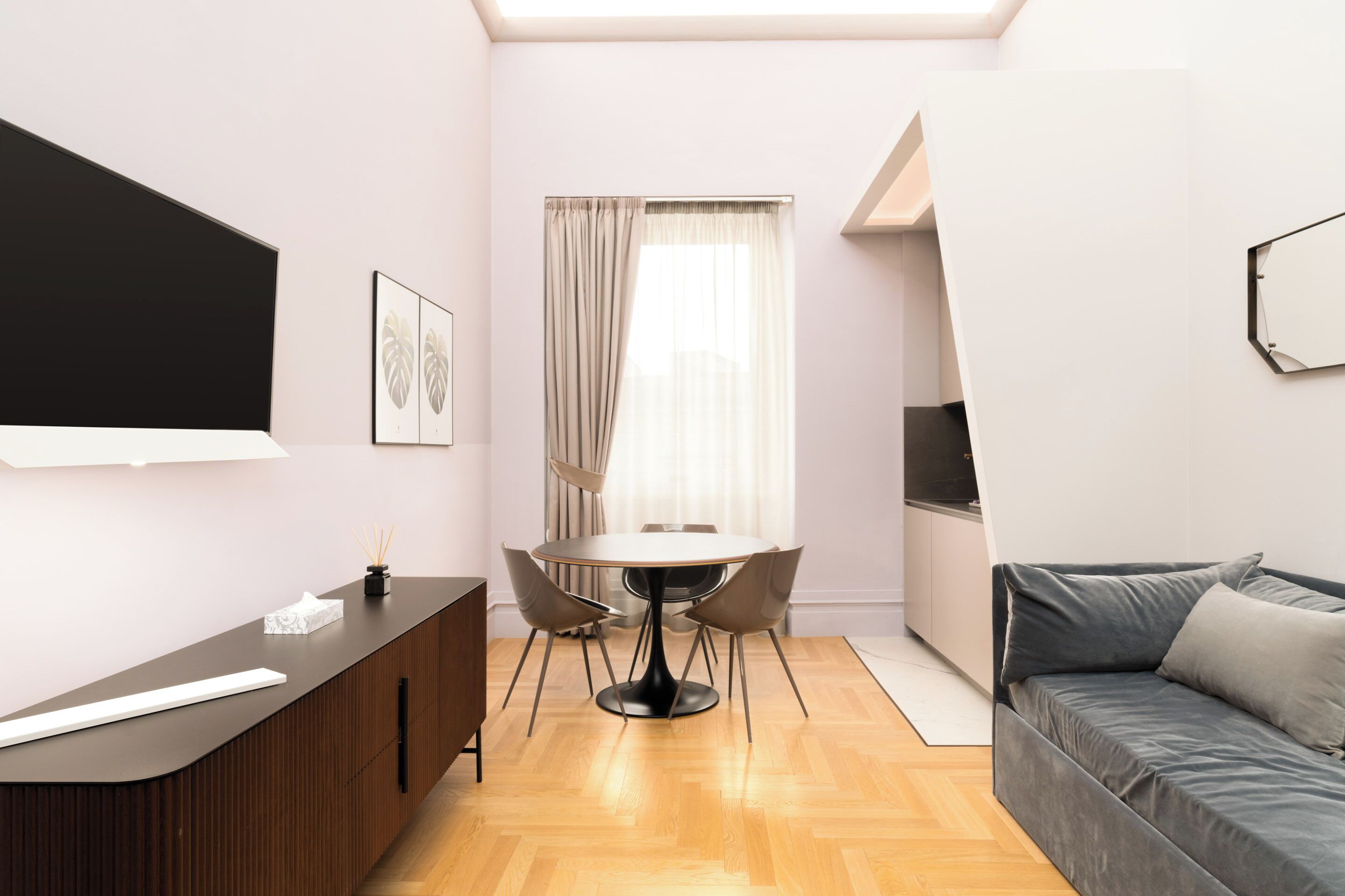 A modern room with a wooden floor, a round dining table with chairs, a sofa, a TV mounted on the wall, and a window with curtains.