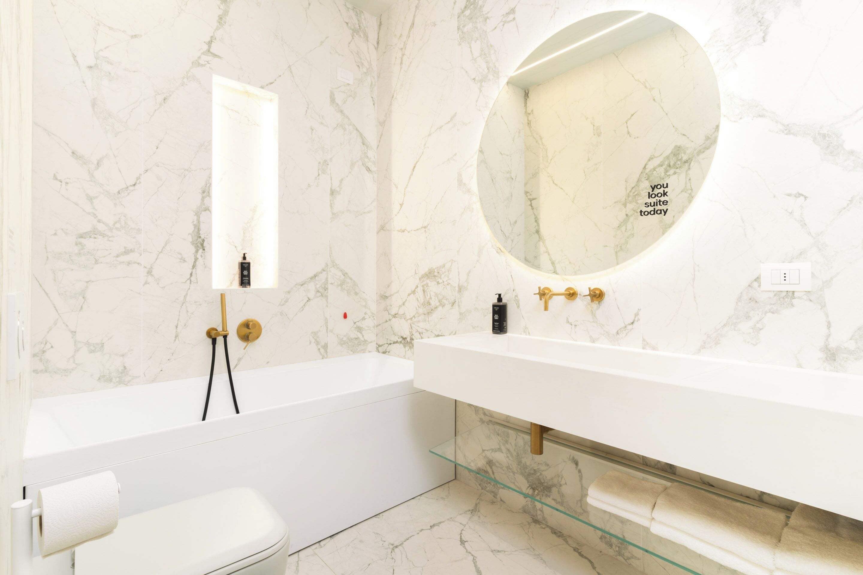 Un baño moderno con pared de mármol, un espejo redondo, una bañera blanca y un estante de vidrio.