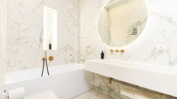 Un baño moderno con pared de mármol, un espejo redondo, una bañera blanca y un estante de vidrio.