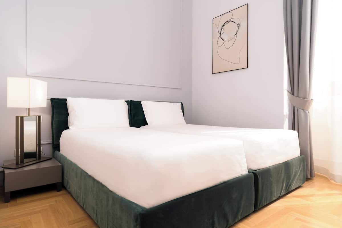 A neatly made double bed with white linens and a green bed skirt, a bedside table with a lamp, and a wall-mounted abstract art piece.