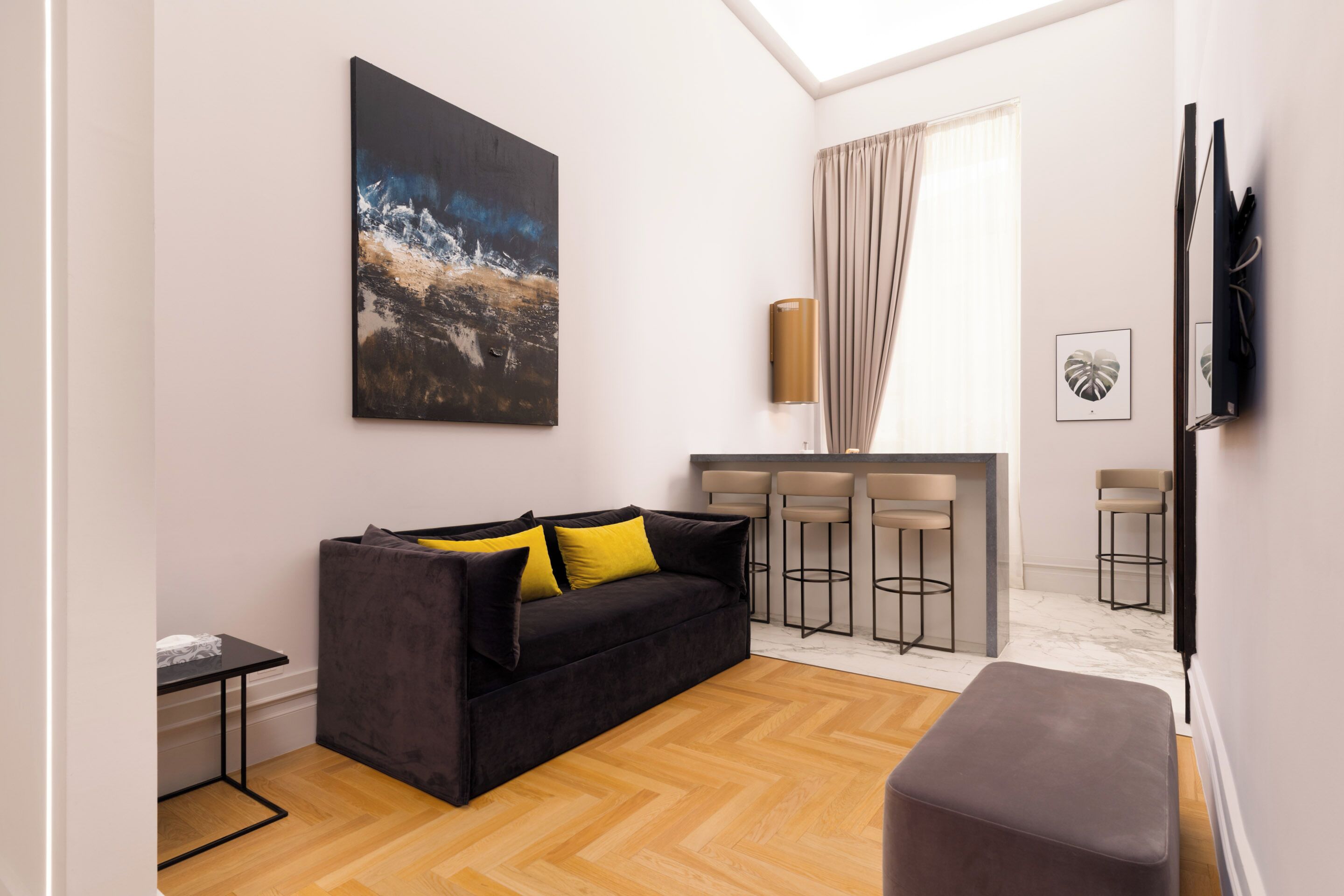 A modern living room with a black sofa, a coffee table, and a large painting on the wall. There are two bar stools and a television mounted on the wall.