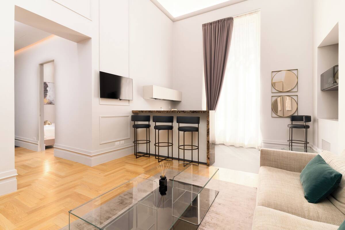 A modern living room with a glass coffee table, a beige sofa, and a kitchen area with bar stools.