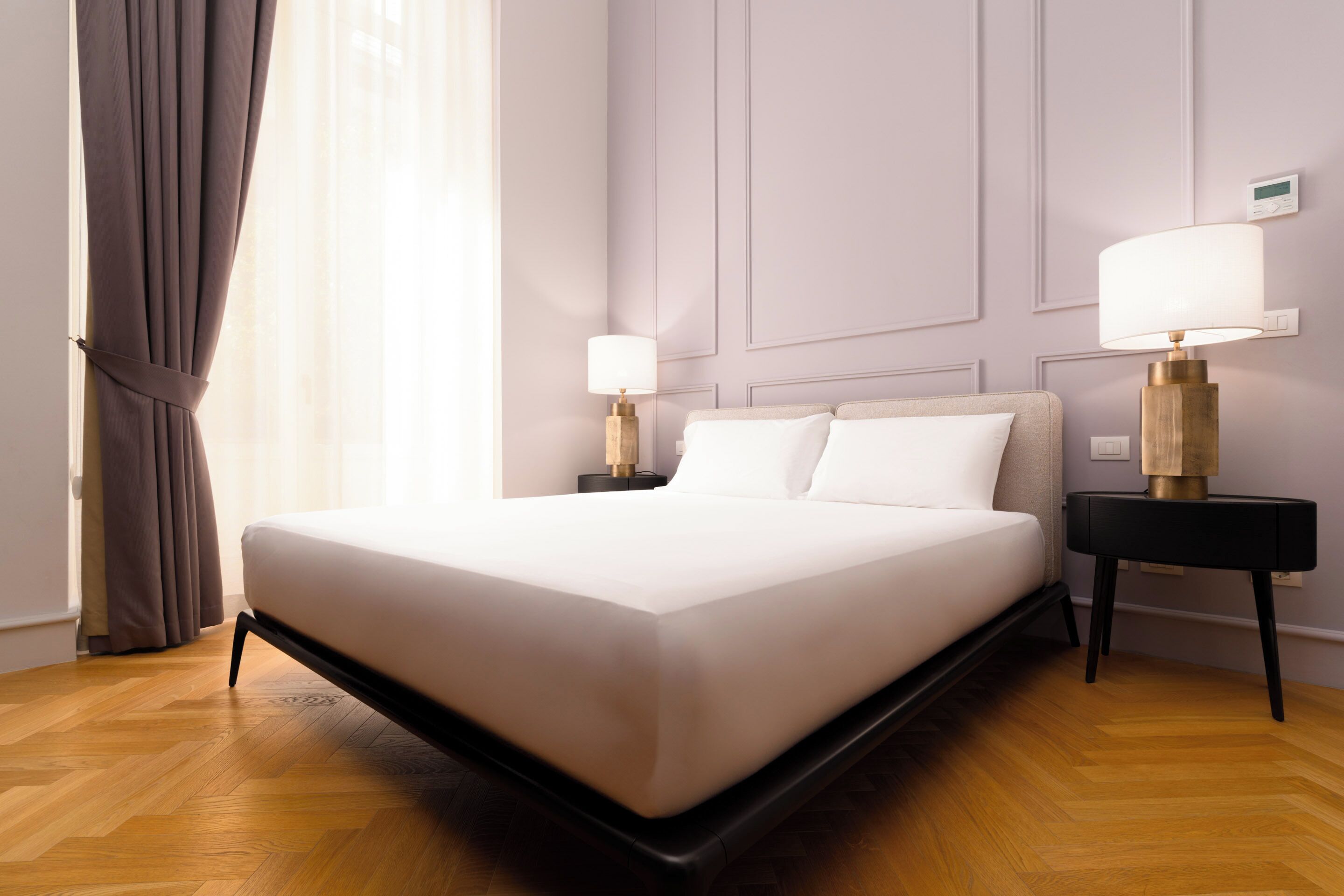 A modern hotel room with a large bed, two bedside tables with lamps, a wooden floor, and a view of the outdoors through sheer curtains.
