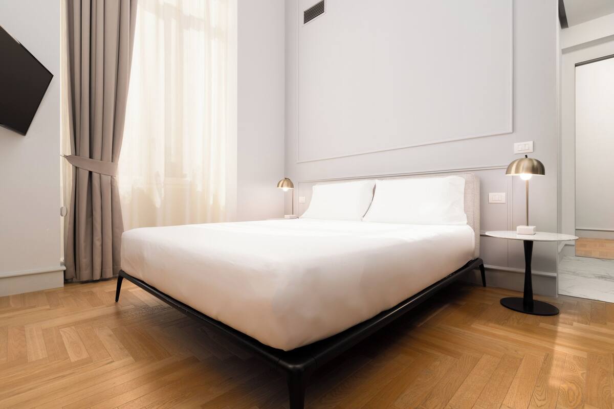 A modern hotel room with a large bed, wooden flooring, a flat-screen TV mounted on the wall, and a small round table with a lamp.